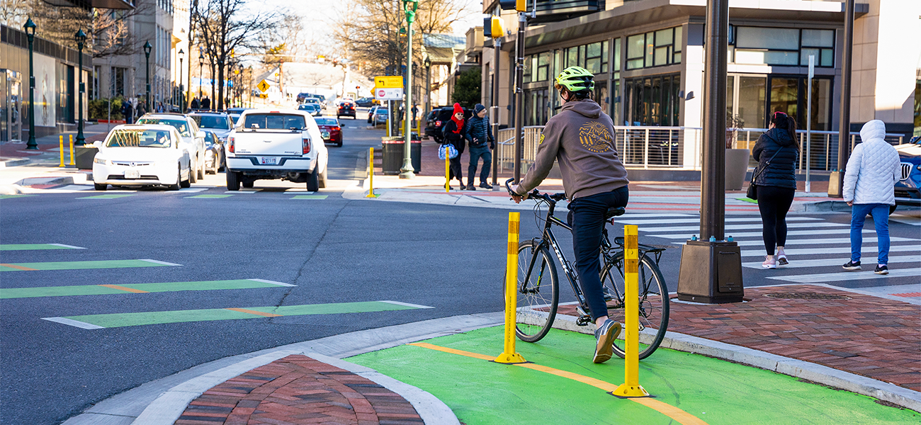A cyclist wearing a helmet rides in a green bike lane at a city intersection, while cars and pedestrians move through the area on a sunny day.