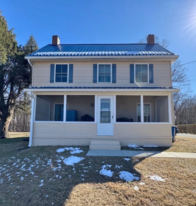 Two-story beige house with blue shutters and a metal roof, featuring a screened front porch. Patchy snow is visible on the ground in front of the house.