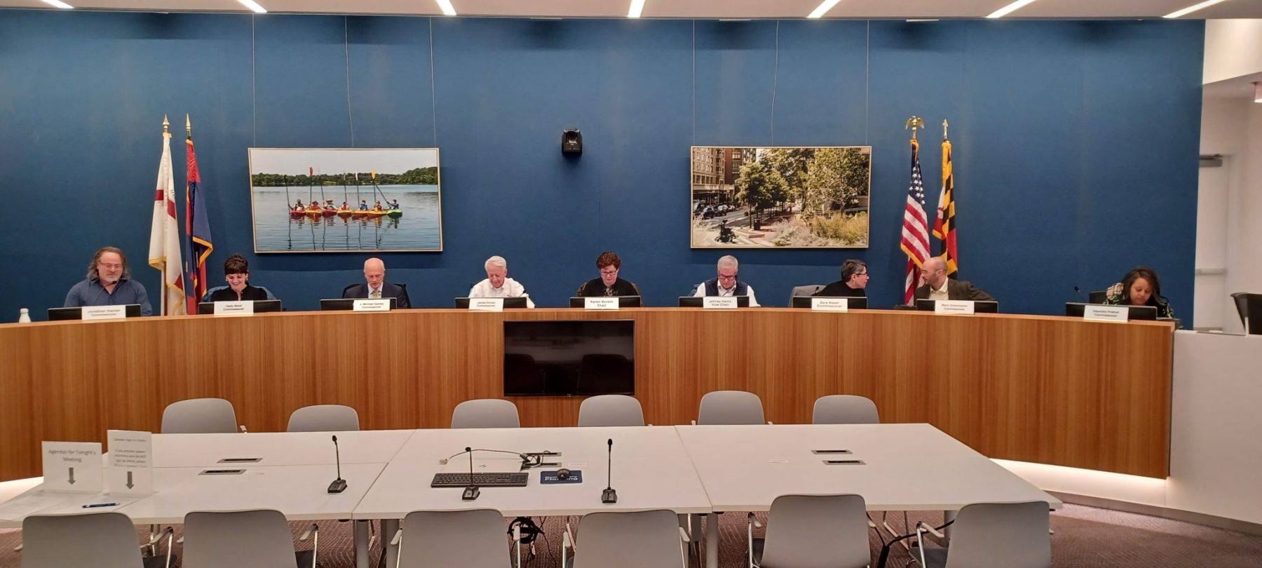 Nine people sit at a curved panel desk in a council meeting room; two flags and framed photos are displayed on a blue wall behind them.