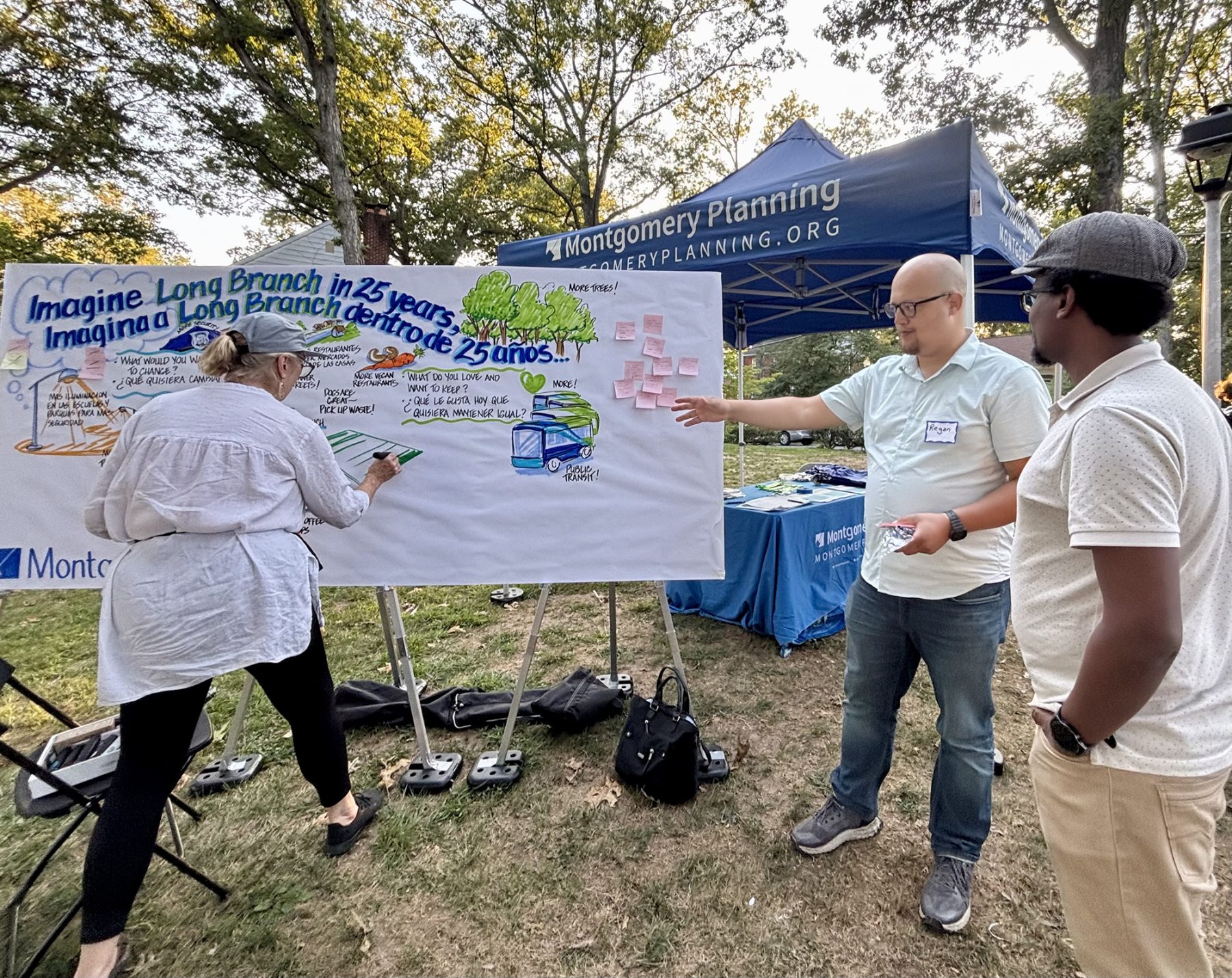 Three people interact with a community planning display outdoors, adding sticky notes and writing ideas on a large board under a Montgomery Planning tent.
