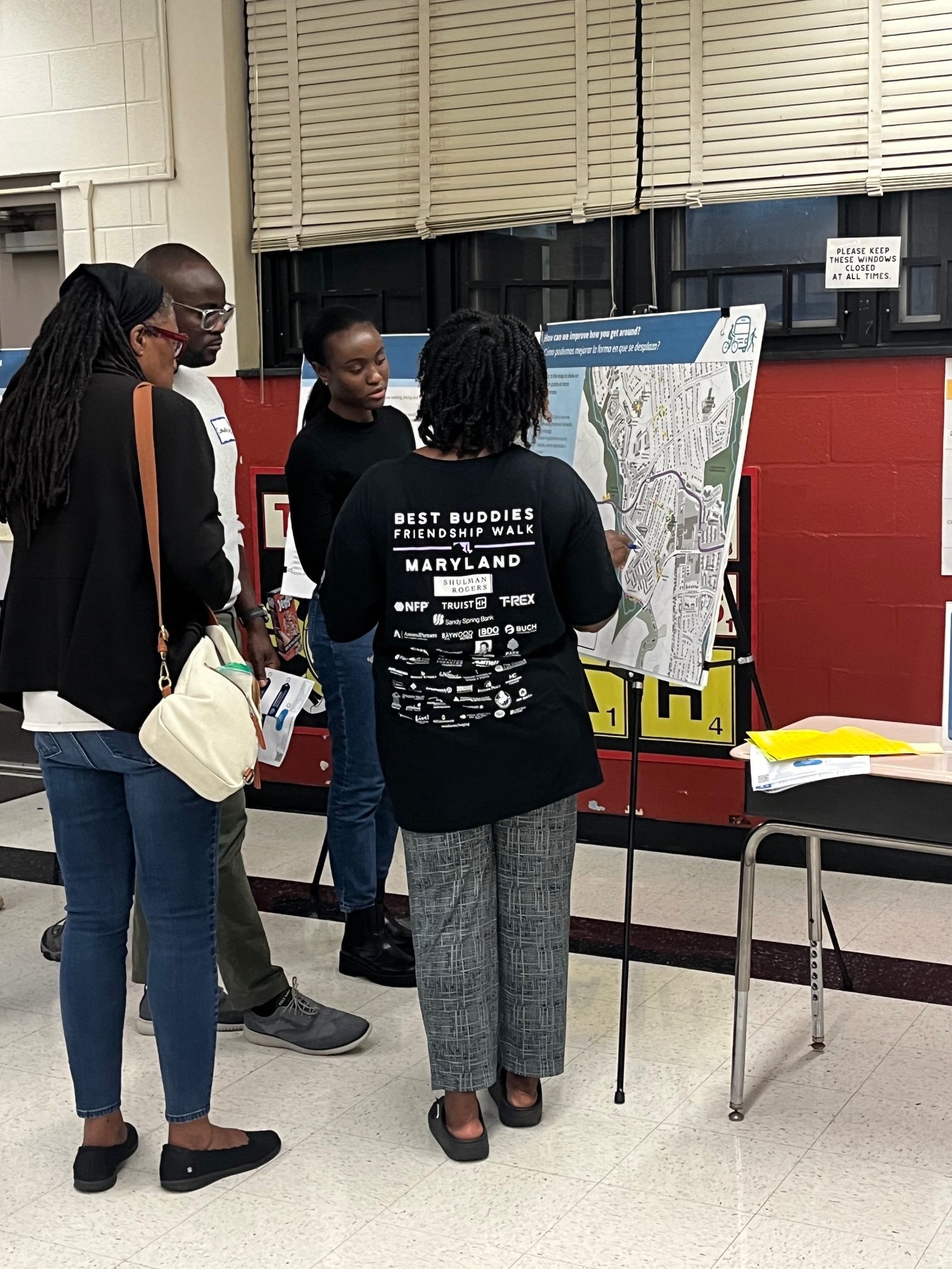 Three people stand around a map display on an easel in an indoor setting, discussing or examining the map.
