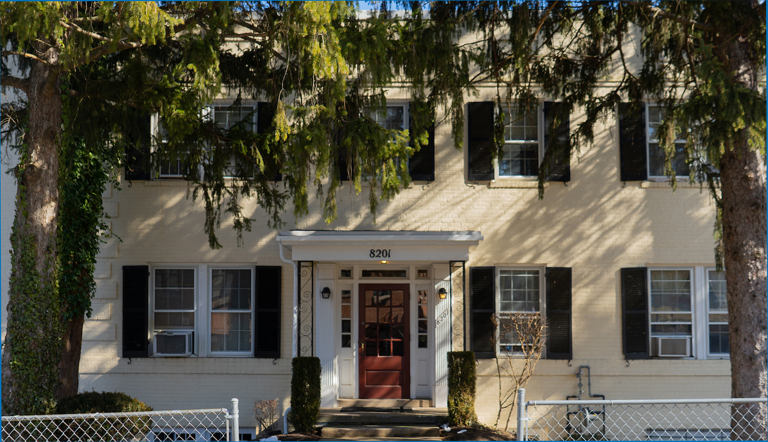 A two-story beige apartment building with black shutters, air conditioning units in windows, and the address 8201 above the entrance, partially shaded by large trees.