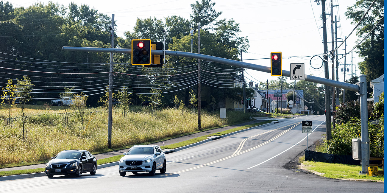 Two cars wait at a red traffic light on a suburban road with a right turn only lane and visible power lines and trees in the background.