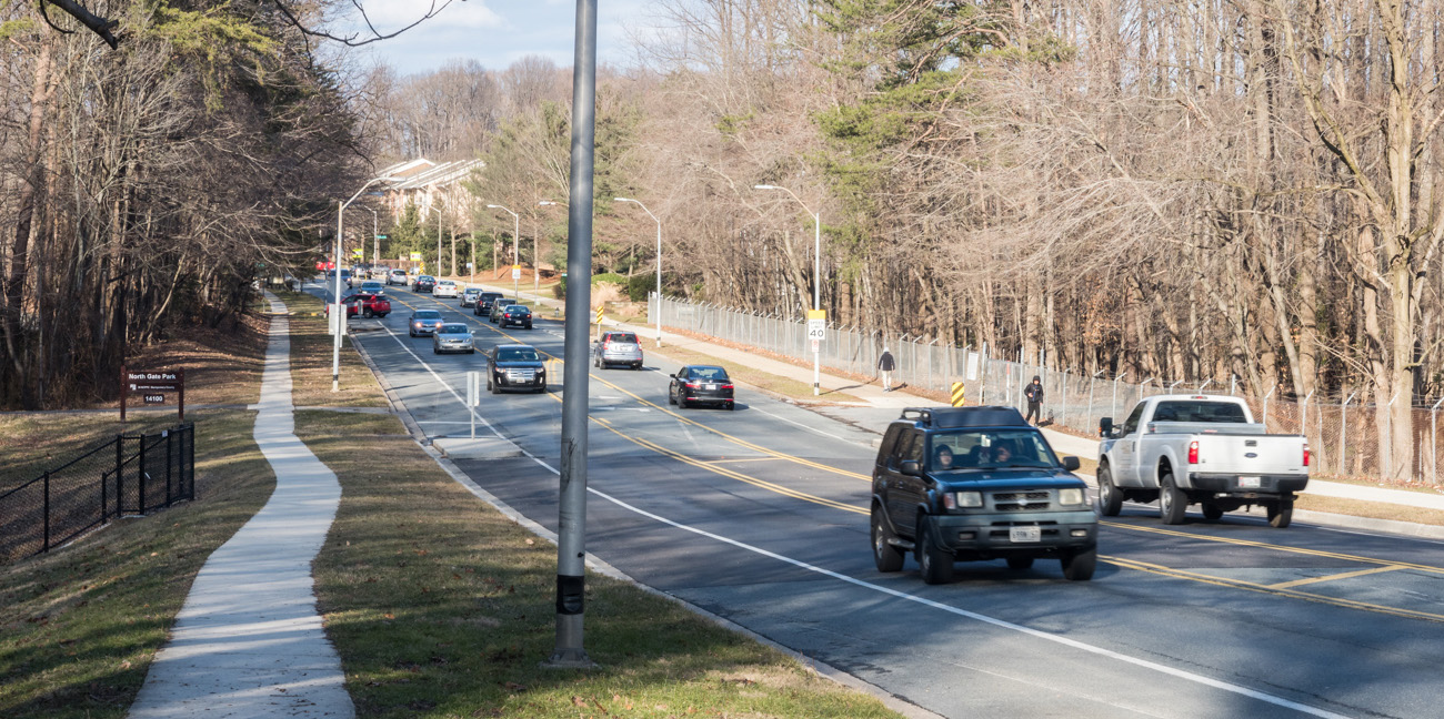 A two-lane road with cars driving in both directions, trees lining the right side, a sidewalk on the left, and a few pedestrians in the distance.