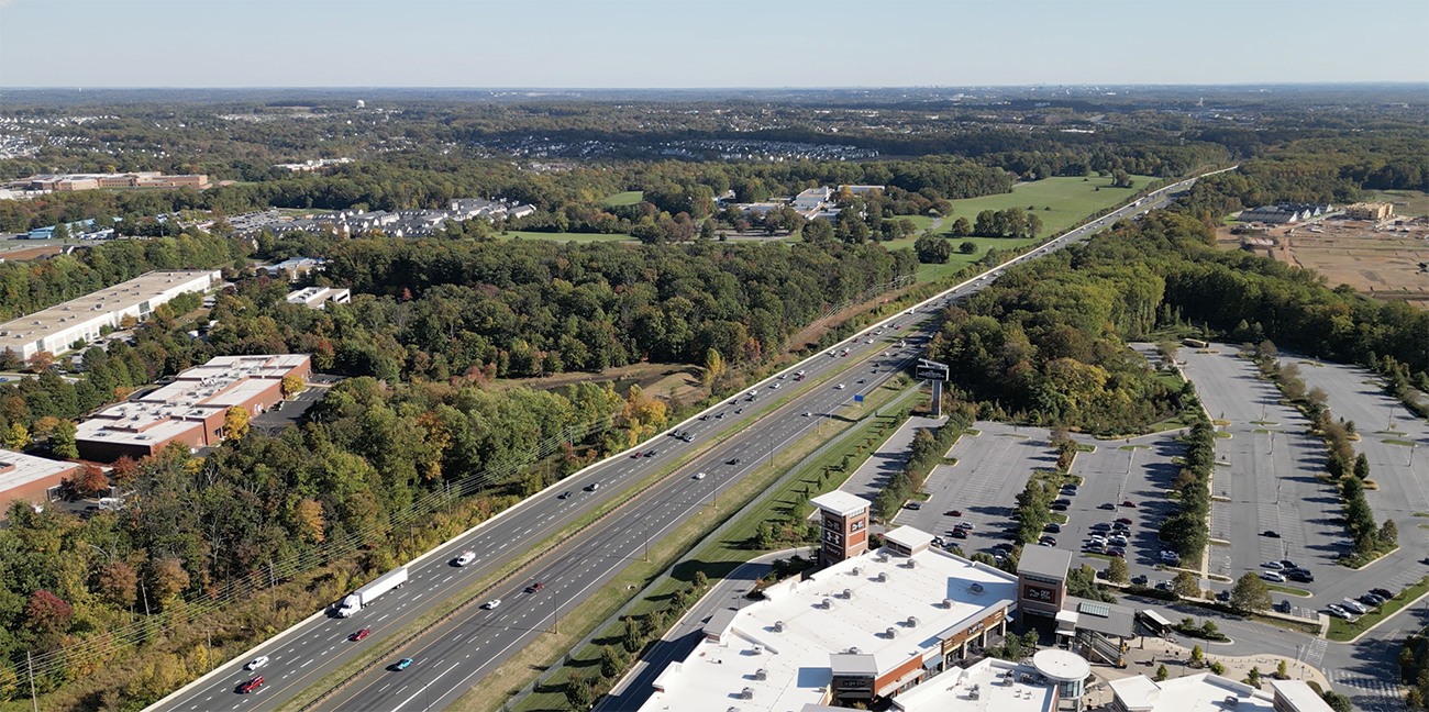 Aerial view of a multi-lane highway running through a green, wooded area, with parking lots, commercial buildings, and scattered vehicles nearby.
