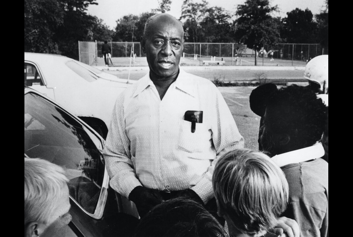 A man stands outdoors near a car, surrounded by children, with trees and a fence visible in the background.