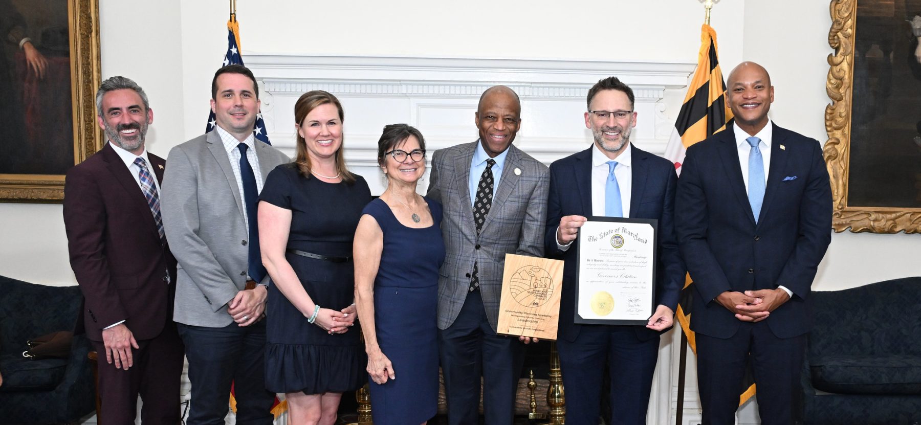 Seven people stand together indoors, two holding awards, with American and Maryland flags and a white fireplace in the background.