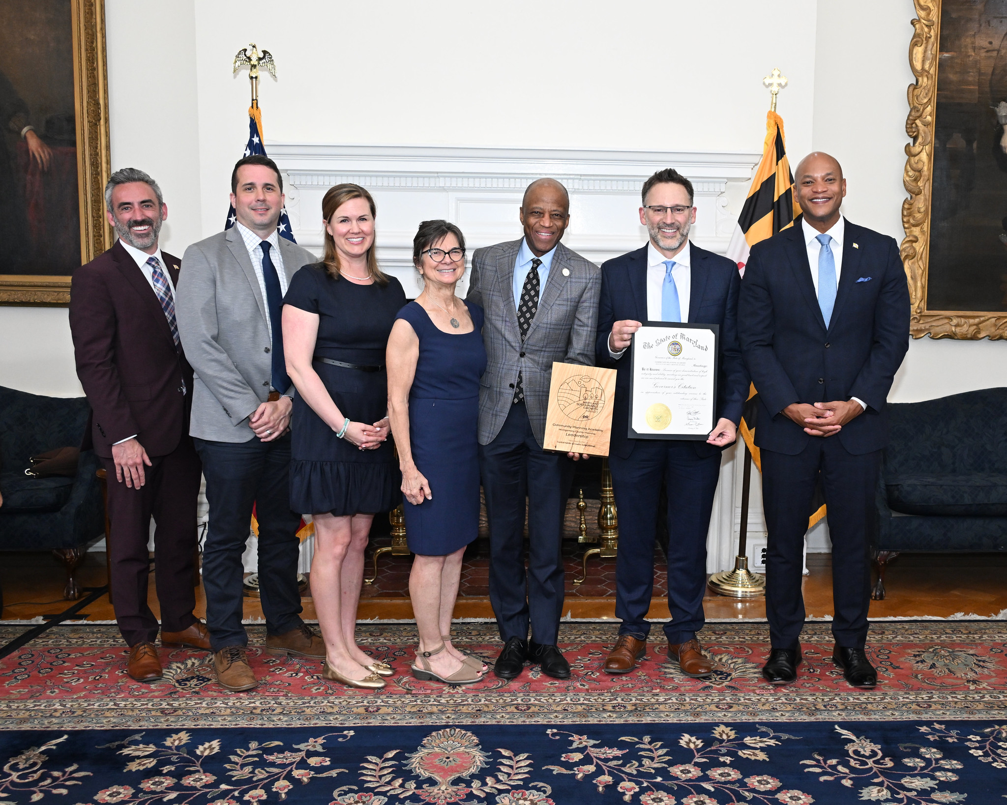 Seven people stand in a formal room, posing for a group photo; one man in the center holds a certificate and plaque. An American flag and Maryland flag are visible in the background.