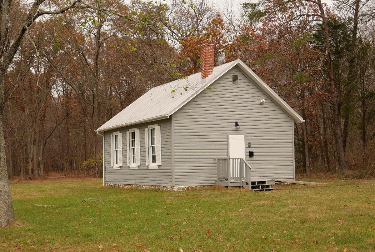 A small, gray, wooden building with a metal roof and brick chimney sits on a grassy lawn, surrounded by trees with autumn foliage.