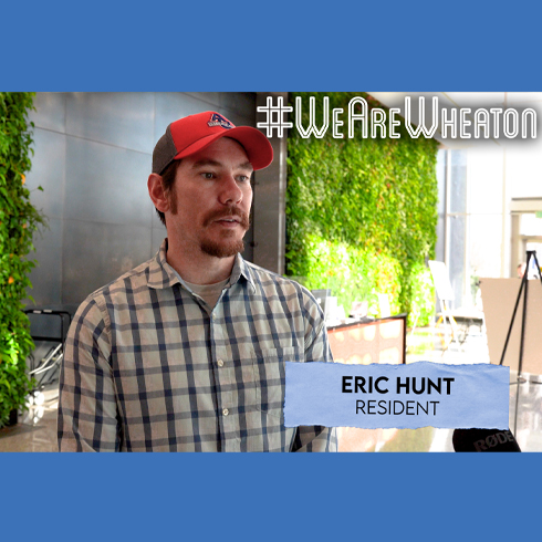 A man in a plaid shirt and red cap stands indoors near a green wall. Text on image: "#WeAreWheaton" and "Eric Hunt, Resident.