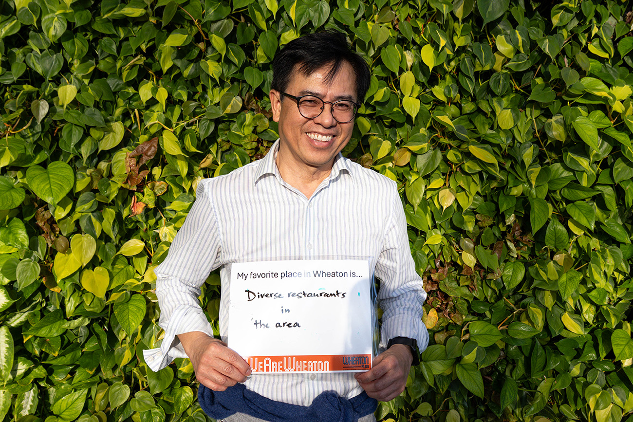 A man stands in front of a leafy green wall, smiling and holding a sign that says, "My favorite place in Wheaton is... Diverse restaurants in the area.