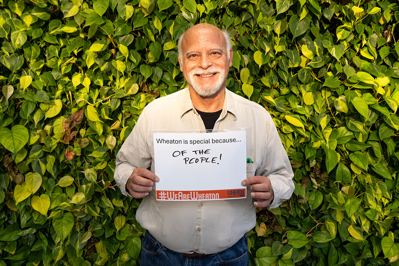 An older man stands in front of a leafy green wall, smiling and holding a sign that reads, "Wheaton is special because... OF THE PEOPLE!" with a #WeAreWheaton hashtag.