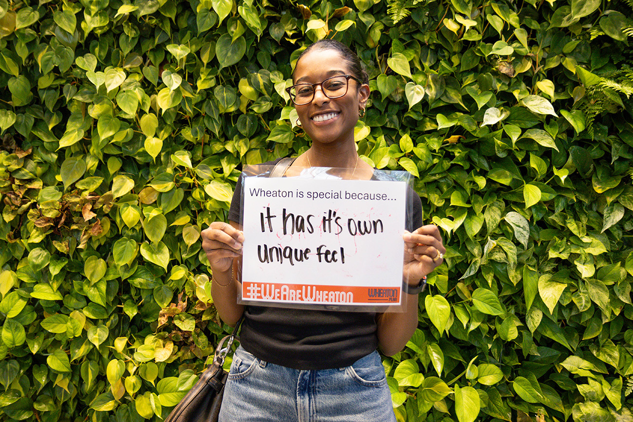 A person stands in front of a green leafy wall, holding a sign that reads, "Wheaton is special because... It has its own unique feel.