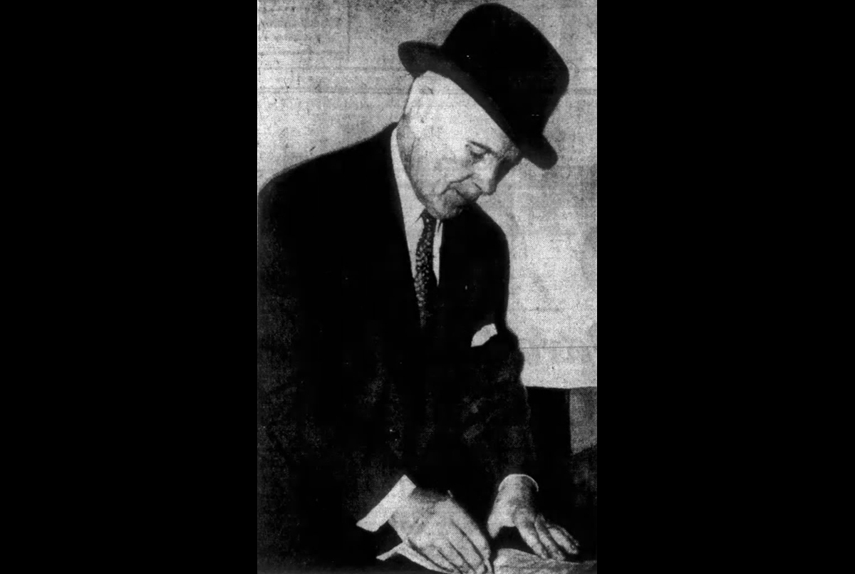 An elderly man in a suit and hat bends over, using his hands to read a document with raised dots, suggesting he is reading braille.