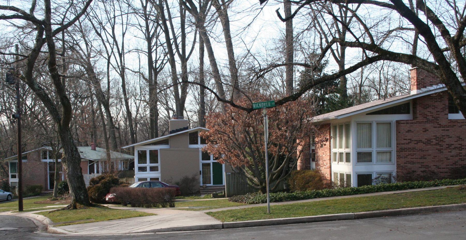 Residential street with mid-century modern houses, bare trees, manicured lawns, and a street sign at the corner under a clear sky.