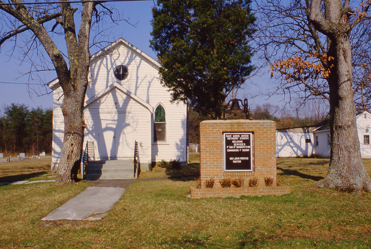 A small white church with arched windows stands behind a brick monument sign and a bell, surrounded by leafless trees and grassy lawn on a clear day.