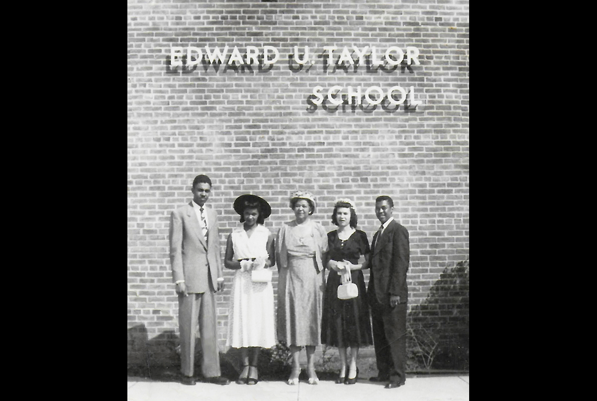 Five people stand in front of a brick wall with the words “Edward U. Taylor School” displayed above them.