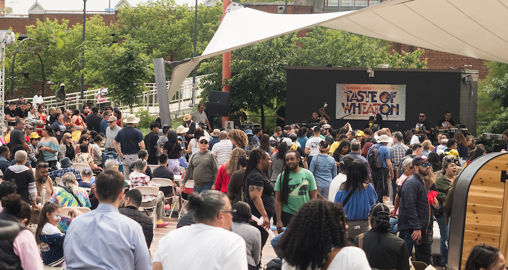 A large crowd gathers outdoors in front of a stage with a "Taste of Wheaton" sign, where musicians are performing. Trees and event tents are visible in the background.