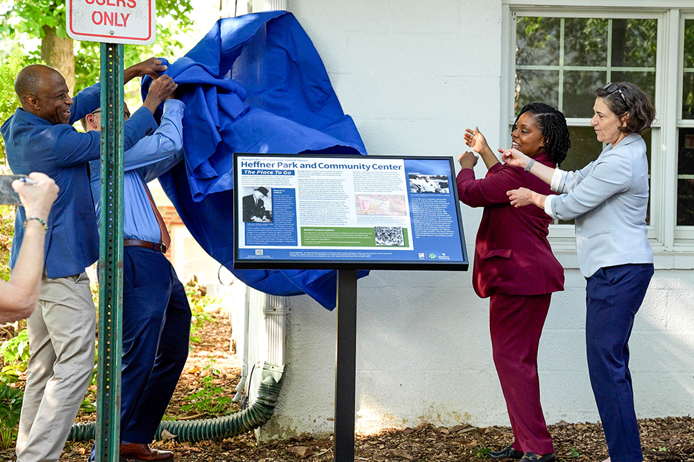 Four people unveil a blue cloth from a new informational sign outside a building labeled "Heffner Park and Community Center.