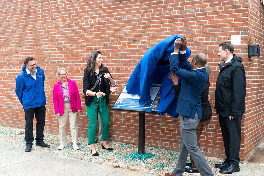 Six people stand by a brick wall as two individuals unveil a blue cloth covering an informational sign or plaque.