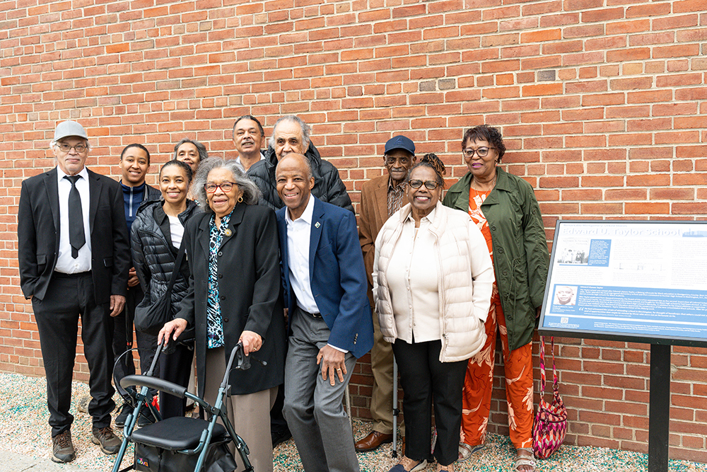 A group of people pose and smile together in front of a brick wall and historical information sign.