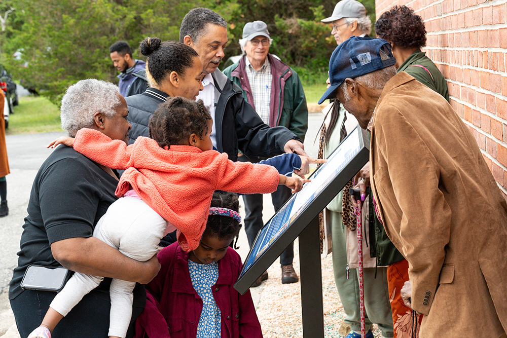 A group of people, including adults and children, gather around and read an outdoor informational sign next to a brick wall.