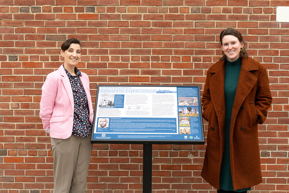 Two women stand on either side of an informational sign titled "Edward U. Taylor School" in front of a brick wall.