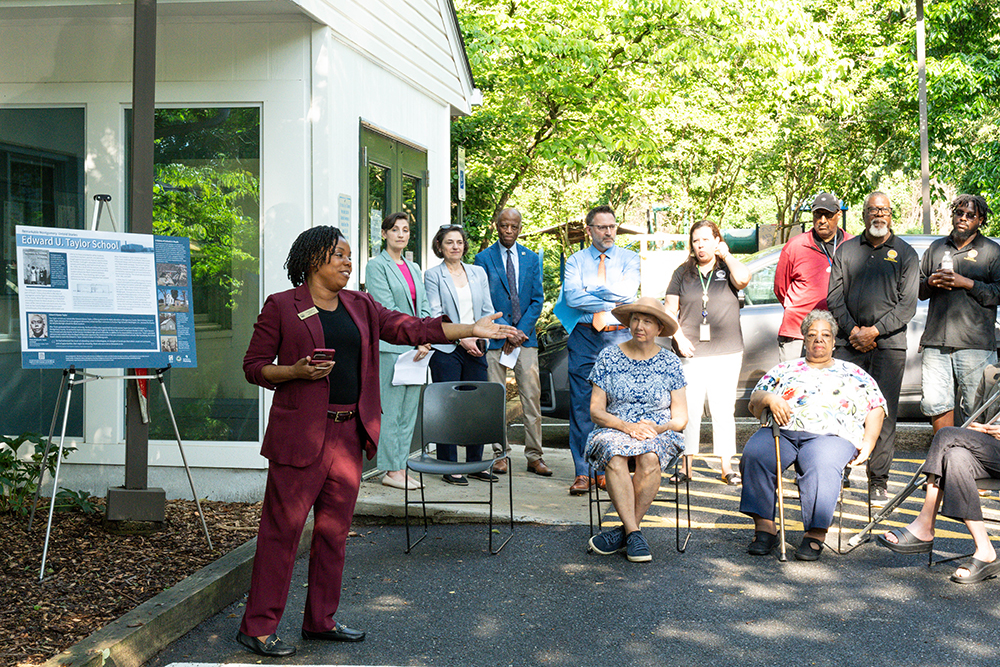 A woman speaks to a seated and standing group outdoors near a building and a display board on an easel.