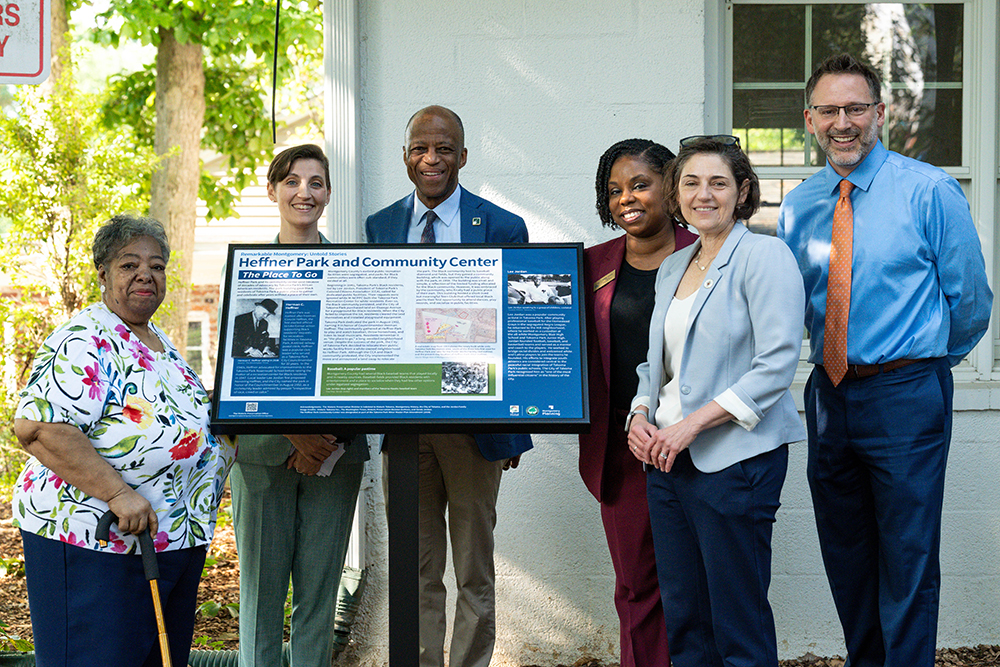 Six adults stand outdoors around an informational sign titled "Hefner Park and Community Center." They are smiling and posing for a group photo beside a light-colored building.