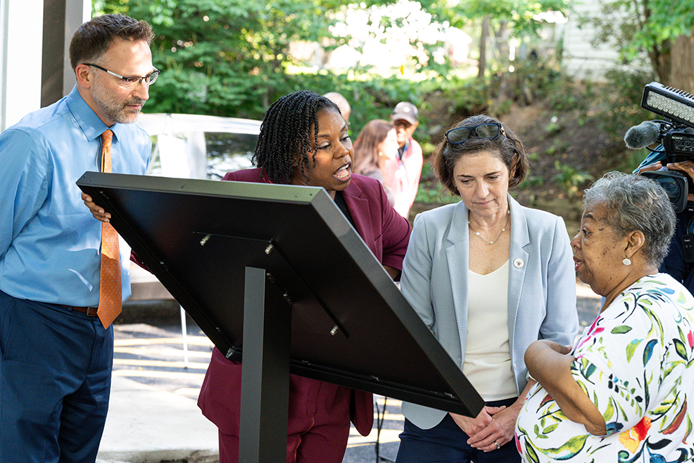 Four adults stand around an outdoor display board, reading and discussing its content, with greenery and other people visible in the background.