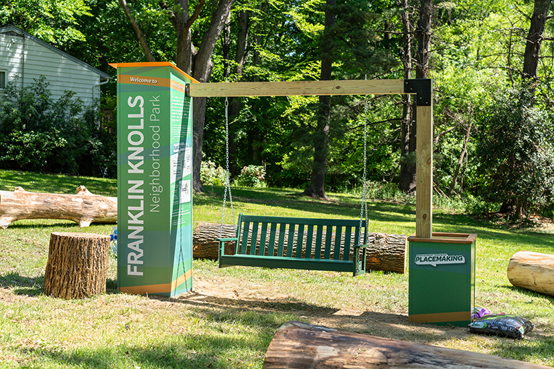 A green swing bench is suspended between two posts with signage reading "Franklin Knolls Neighborhood Park" in a wooded outdoor area.
