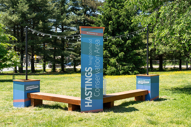 A wooden bench with three blue pillars marked “Hastings Neighborhood Conservation Area” stands in a grassy park surrounded by trees.