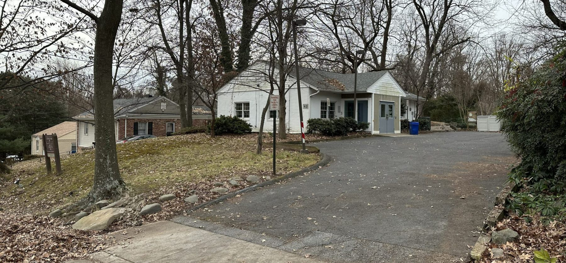 A small white house with a gray roof sits at the end of a paved driveway, surrounded by leafless trees and fallen leaves. A blue recycling bin is near the entrance.