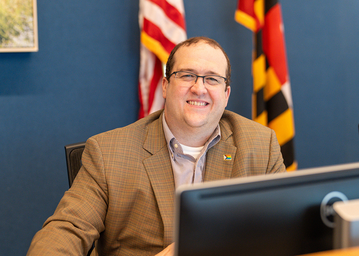 A man in a suit jacket sits at a desk in front of a computer, smiling, with American and Maryland flags and a Montgomery Planning logo in the background.