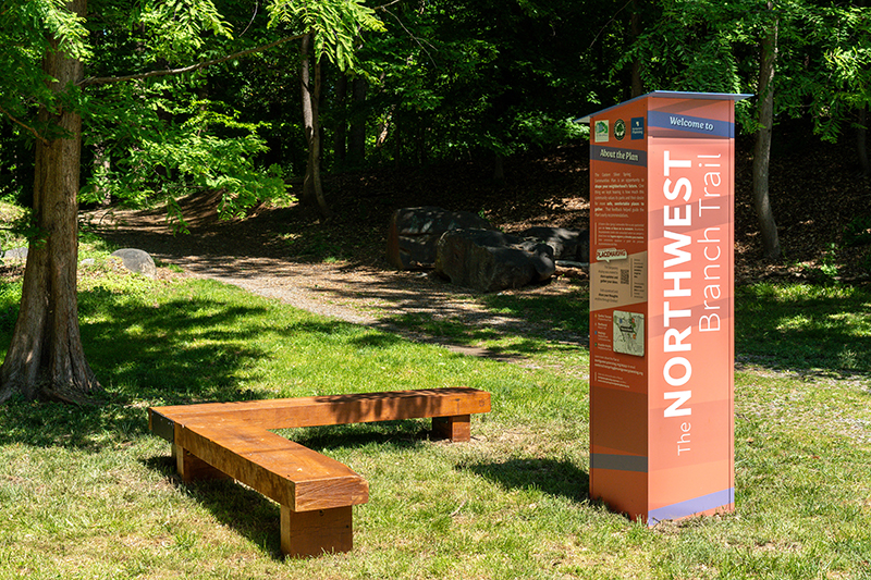 A wooden bench and informational sign for the Northwest Branch Trail are situated on a grassy area beside a shaded forest path.