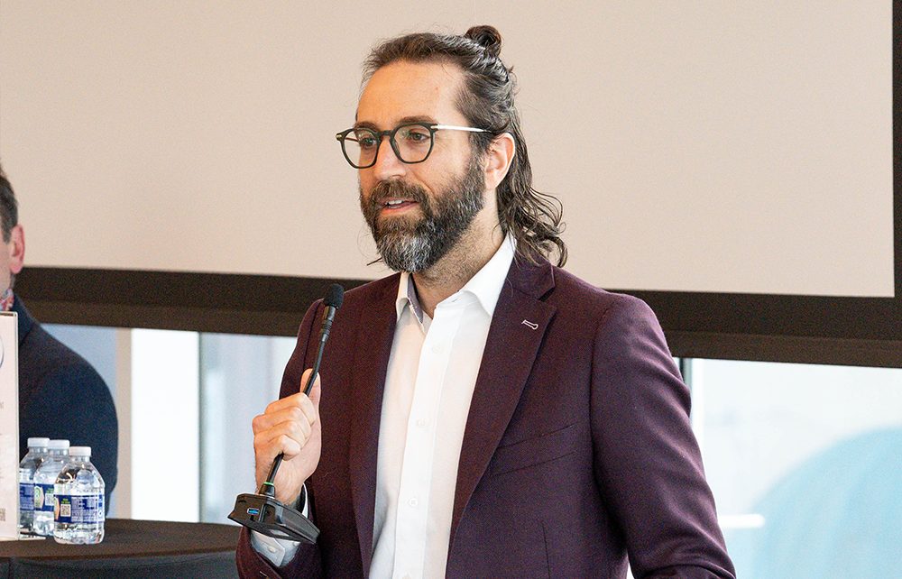 A man with glasses and a beard, wearing a maroon blazer and white shirt, speaks into a microphone at an indoor Montgomery Planning event. Bottled water and papers are visible on the table nearby.