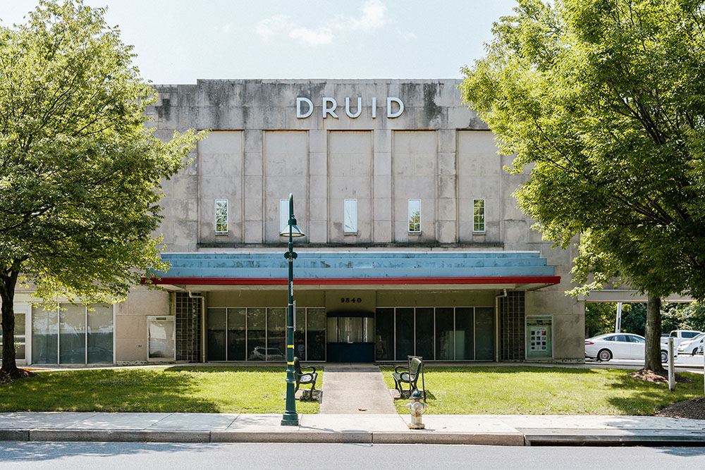 A concrete building with "DRUID" on the facade, large windows, and a covered entrance; two benches and a lamppost are in the grassy area in front, with trees on both sides.