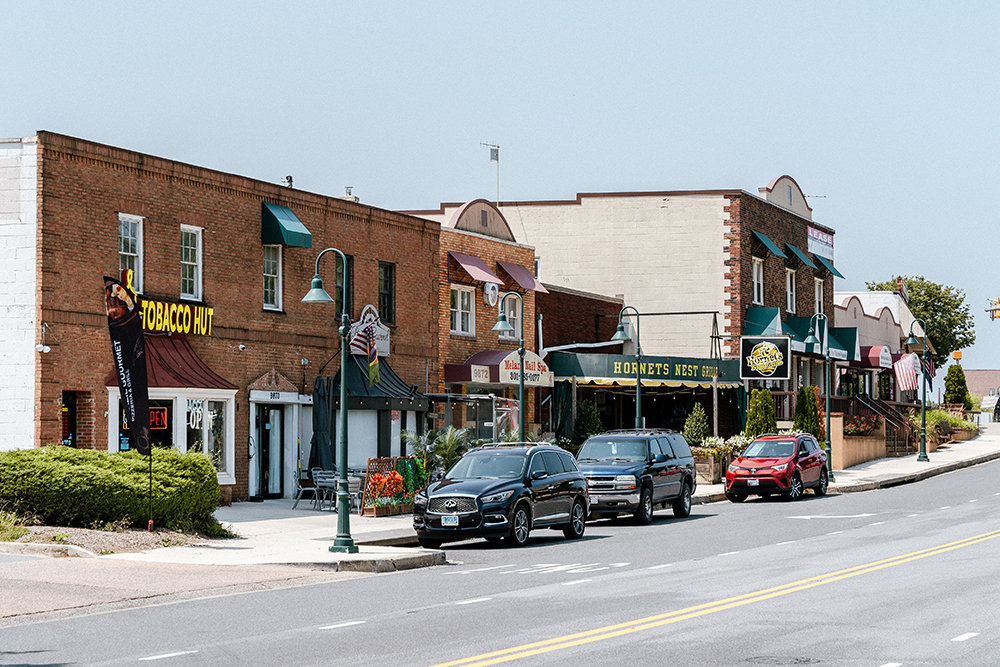 A row of brick storefronts and restaurants with awnings, parked cars along the street, and clear skies overhead.