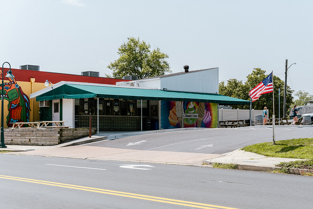 Single-story diner with green awning, outdoor seating, mural, and an American flag displayed; clear blue sky and light traffic.