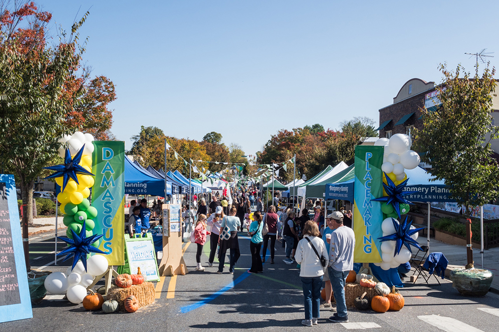 People walk through an outdoor street fair lined with blue tents, balloons, pumpkins, and two vertical banners reading "Damascus" and "Placemaking" on a sunny day.