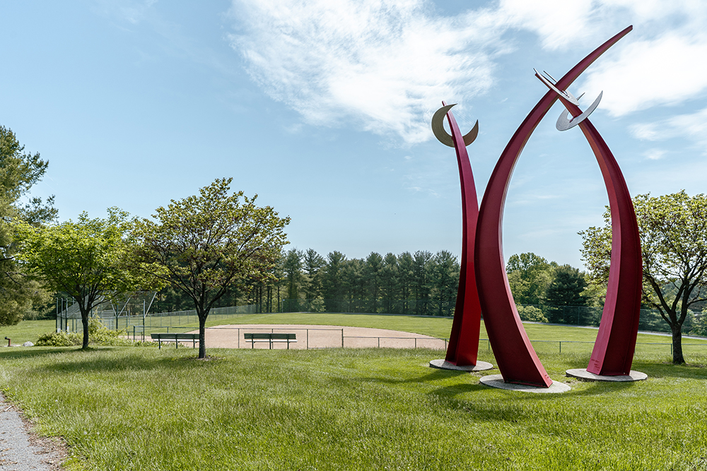 Large red metal sculpture stands on grass near trees, with a baseball field and benches visible in the background under a partly cloudy sky.