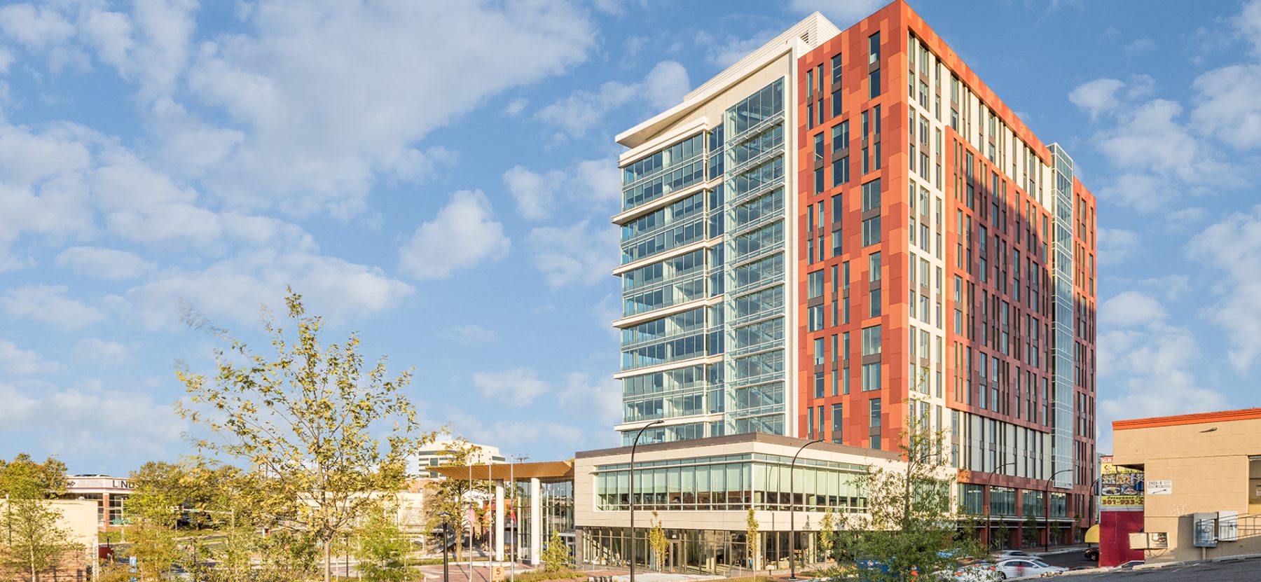 A modern, multi-story office building with glass and orange exterior panels stands against a partly cloudy sky, surrounded by trees and adjacent structures.