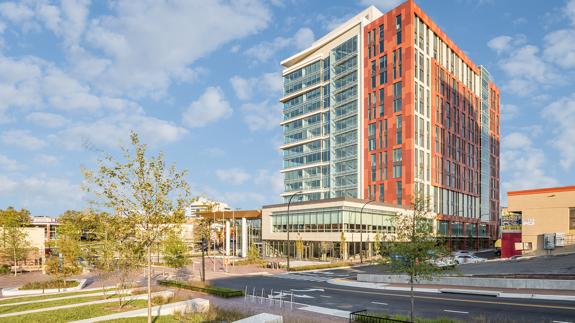 A modern multi-story building with glass and red panels stands on a city street corner, surrounded by trees and a landscaped public area under a partly cloudy sky.