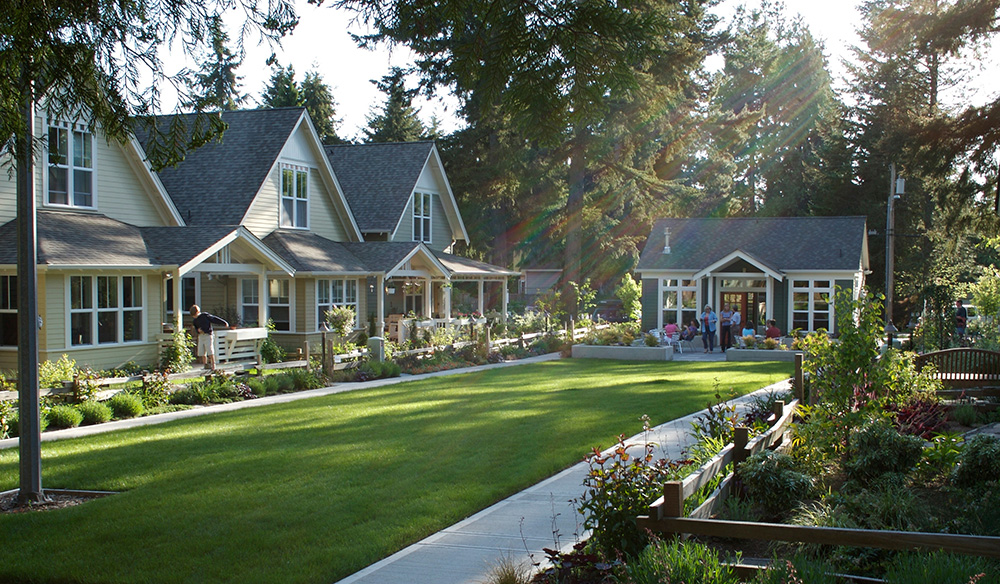 A row of yellow suburban houses with porches faces a well-kept green lawn and gardens, thoughtfully designed in line with Montgomery Planning principles, bordered by a sidewalk on a sunny day with trees in the background.